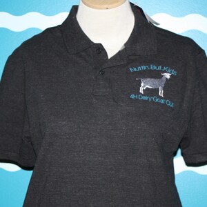 K&ouml;nnte beinhalten: Ein dunkelgraues Poloshirt mit einem blauen gestickten Logo, auf dem "Nuttin But Kids 4H Dairy Goat Out" steht, und einem kleinen Bild einer Ziege.