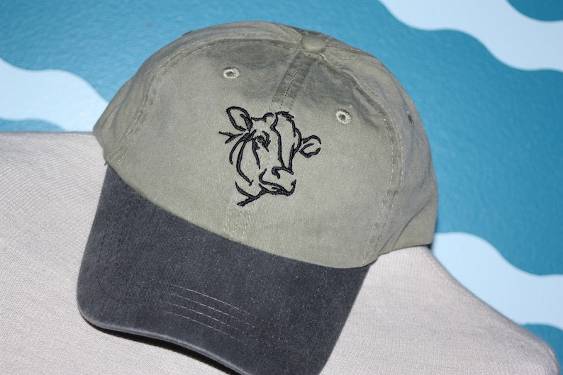 May include: A green and black baseball cap with a black embroidered cow head design.