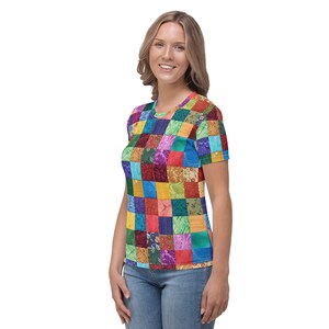 May include: A woman is wearing a short-sleeved t-shirt with a colourful patchwork design. The shirt is made up of squares of different colours and patterns, including red, blue, green, yellow, and purple.