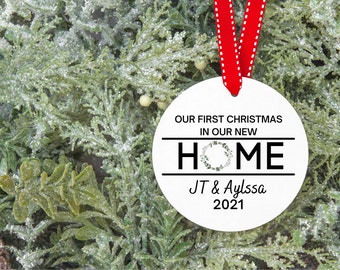 May include: A white Christmas ornament with the text "OUR FIRST CHRISTMAS IN OUR NEW HOME" and the names "JT & Aylssa 2021". The ornament hangs from a red ribbon and is set against a backdrop of green and white frosted foliage.