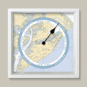 May include: Square white-framed tide clock with a nautical chart design. The clock face has a light blue ring indicating tide times, with black hands. The chart shows coastal details. The clock is in a white frame.