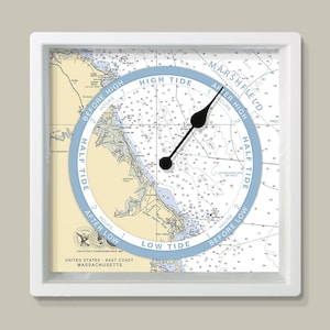 May include: Square white-framed tide clock with a nautical chart of the Massachusetts coast. The clock face has a light blue ring indicating tide times, with black hands showing the current tide phase. The chart includes text such as "High Tide" and "Low Tide".