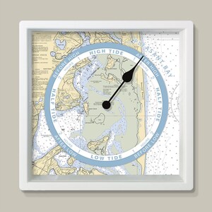 May include: A square white-framed tide clock. The clock face features a nautical chart with a blue and white tide dial. Black clock hands indicate the time relative to high and low tide. The dial reads "High Tide" and "Low Tide".