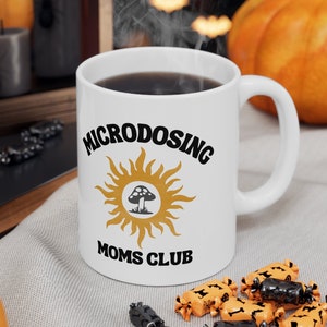 May include: White ceramic coffee mug with black text that reads "Microdosing Moms Club" and a yellow sun with a black mushroom in the center. The mug is sitting on a white surface with orange and black candy.