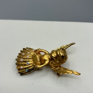Pu&ograve; includere: Broche a forma di angelo in oro con dettagli in strass trasparenti. L'angelo tiene un piccolo libro tra le mani e ha le ali spiegate.