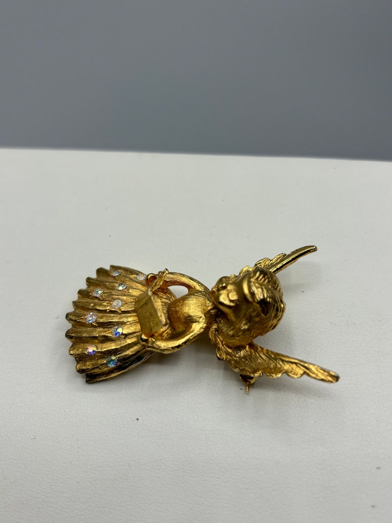 Pu&ograve; includere: Broche a forma di angelo in oro con dettagli in strass trasparenti. L'angelo tiene un piccolo libro tra le mani e ha le ali spiegate.