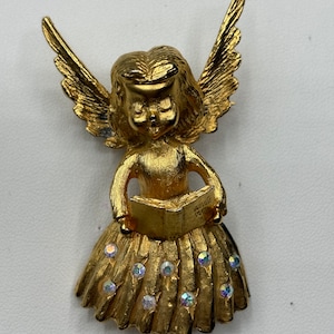 Pu&ograve; includere: Una statuetta di angelo dorata con ali, che tiene un libro e indossa una gonna con strass iridescenti.