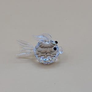 May include: A clear, faceted crystal figurine shaped like a small, round fish. The body features a geometric, diamond-cut pattern that reflects light. It has a smooth, transparent tail, a dorsal fin, and two small, dark, circular eyes on its face.