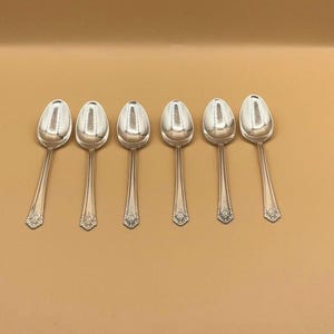 May include: Six silver spoons displayed in a row. Each spoon features a rounded bowl and a long, slender handle with a decorative floral design at the base. The spoons are set against a plain, light brown background.