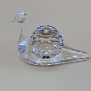 May include: A clear glass figurine shaped like a snail with a smooth, elongated body and a faceted, spherical shell that reflects light. The piece features a curved neck and a small, pointed head, crafted from transparent material throughout.