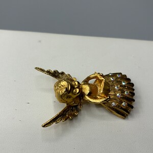 Pu&ograve; includere: Broche a forma di angelo in oro con accenti di strass trasparenti. L'angelo &egrave; seduto con le ali spiegate e tiene un piccolo oggetto tra le mani.