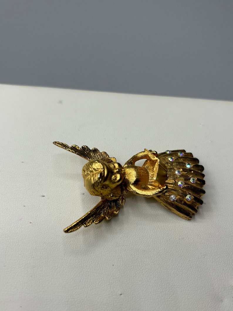 Pu&ograve; includere: Broche a forma di angelo in oro con accenti di strass trasparenti. L'angelo &egrave; seduto con le ali spiegate e tiene un piccolo oggetto tra le mani.