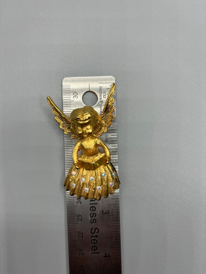 Pu&ograve; includere: Figurina di angelo dorata con ali, che tiene un libro e indossa un vestito con strass. La figurina &egrave; alta circa 2,5 cm.