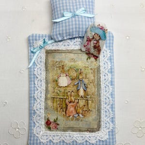 May include: A Beatrix Potter dollhouse baby nursery set featuring a quilt, pillow, and bunny doll. The quilt has a blue and white gingham pattern with a framed illustration of Peter Rabbit and his family. The set includes a small pillow and a bunny doll.