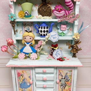 May include: A miniature Alice in Wonderland themed cabinet painted in light blue and pink. The cabinet features various character figurines, a clock, and the word "Wonderland". The cabinet has drawers and doors with Alice illustrations.