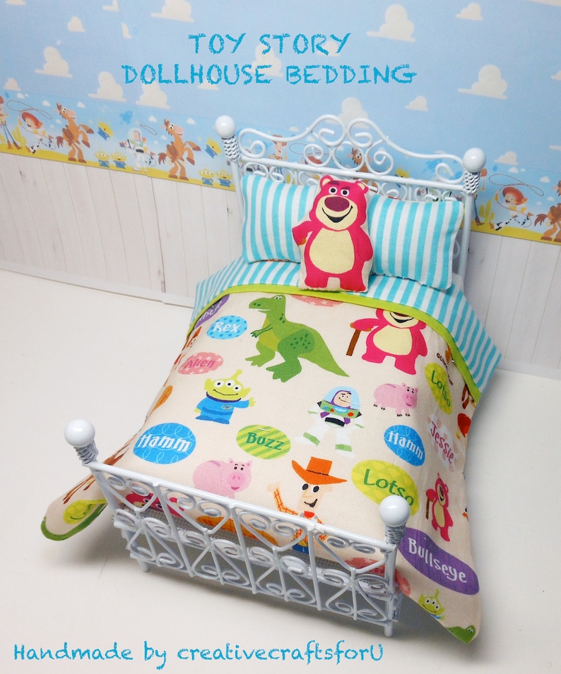 May include: White dollhouse bed with a white metal frame and a Toy Story themed bedding set. The bedding features a white background with colorful characters from the movie, including Buzz Lightyear, Woody, and Jessie. The pillow is pink with a smiling bear face.