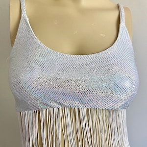 May include: A white, shimmering crop top with thin straps and a scoop neckline. The top is covered in small, reflective sequins and features a fringe detail along the bottom edge. The fringe is made of long, white strands.