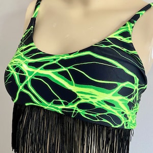 May include: Black crop top with neon green lightning bolt design and black fringe. The top has thin straps and a scoop neckline. The fringe hangs from the bottom hem of the top. The fabric is a stretchy material.