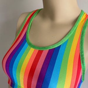 Puede incluir: Un sujetador deportivo a rayas arcoíris con ribete verde. El sujetador presenta rayas verticales en varios colores, incluyendo rojo, naranja, amarillo, verde, azul y morado. El escote y las correas están delineados con una tela verde brillante.