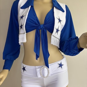 May include: A blue and white two-piece outfit. The top is a cropped, long-sleeved shirt with a tie front. The white accents feature star embellishments and fringe. The matching shorts have star details and a rhinestone buckle.
