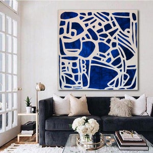 May include: A blue and white abstract painting hangs above a dark blue sofa with white and beige pillows. A glass coffee table sits in front of the sofa with a vase of white flowers and a stack of books.