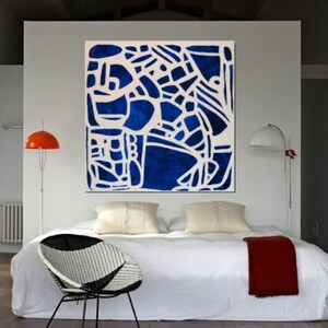 May include: A white bed with a blue and white abstract painting hanging above it. The bed has a white duvet and two white pillows. There is a red blanket on the bed and a black and white wicker chair in front of the bed.