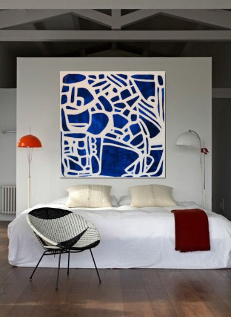 May include: A white bed with a blue and white abstract painting hanging above it. The bed has a white duvet and two white pillows. There is a red blanket on the bed and a black and white wicker chair in front of the bed.