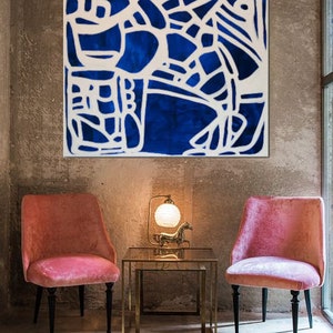 May include: A blue and white abstract painting with a geometric design hangs on a concrete wall above two pink velvet chairs and a gold metal table.