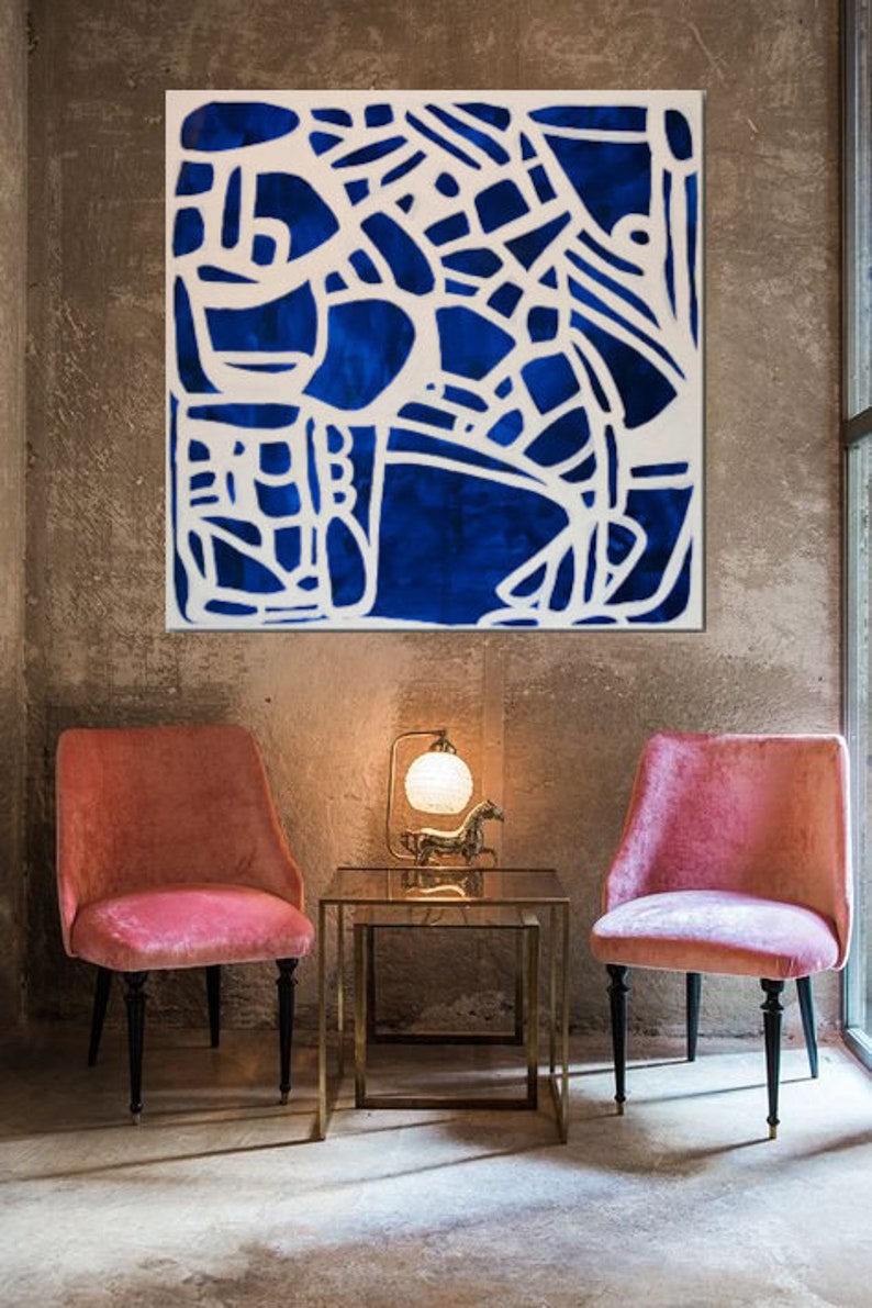 May include: A blue and white abstract painting with a geometric design hangs on a concrete wall above two pink velvet chairs and a gold metal table.