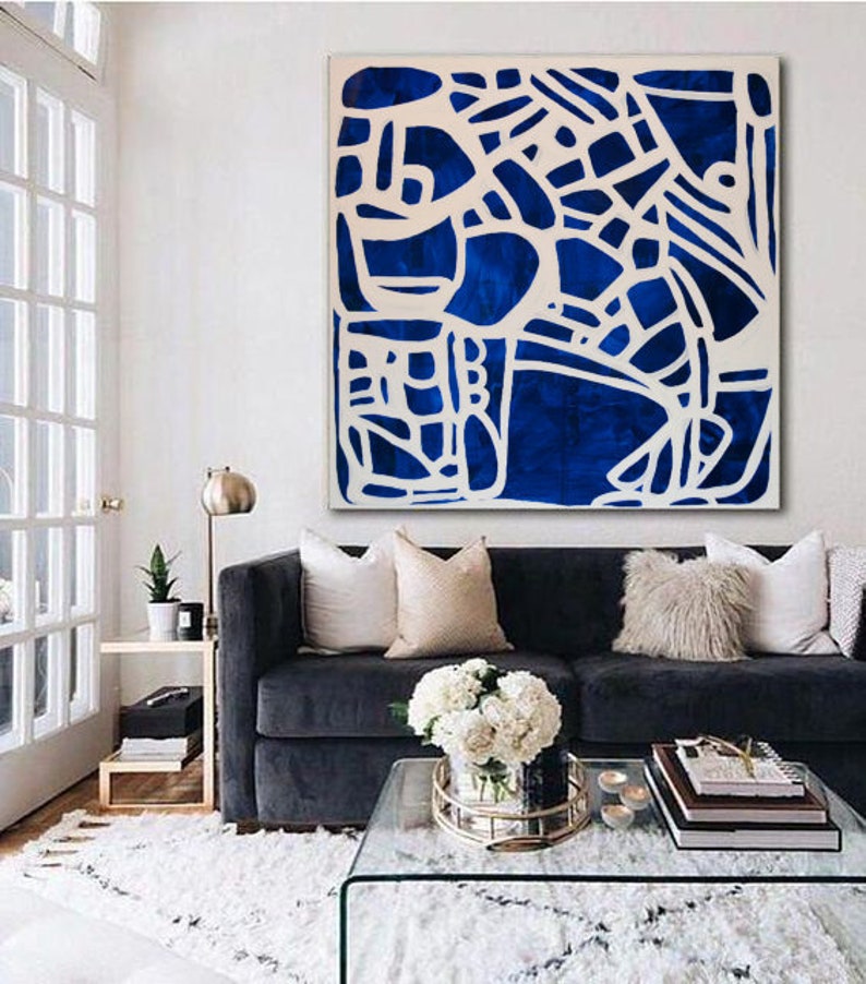 May include: A blue and white abstract painting hangs above a dark blue sofa with white and beige pillows. A glass coffee table sits in front of the sofa with a vase of white flowers and a stack of books.