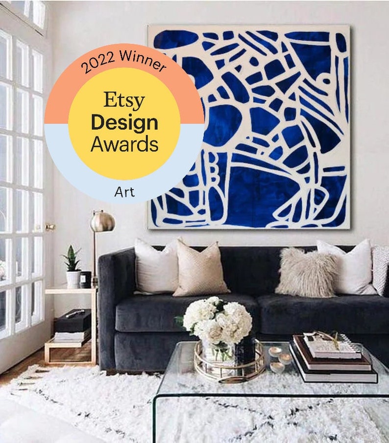 May include: A blue and white abstract art print with a white frame hanging above a dark gray couch with white and beige pillows. The couch is in a living room with a glass coffee table, a white rug, and a gold floor lamp. The art print has a sticker on it that says "2022 Winner Etsy Design Awards Art".