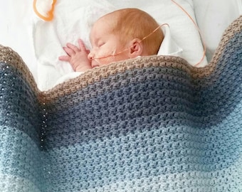 May include: A sleeping infant is nestled under a handmade crochet blanket. The blanket features a gradient of colors, transitioning from brown to blue and white. The baby has red hair and is connected to medical equipment.