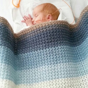 Peut inclure: Un nourrisson endormi est niché sous une couverture crochetée à la main. La couverture présente un dégradé de couleurs, passant du marron au bleu et au blanc. Le bébé a les cheveux roux et est relié à un équipement médical.