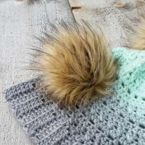 以下が含まれることがあります： グレーとミントグリーンのクロシェ編みビーニーに、茶色のフェイクファーのポンポンが付いている。ビーニーには、「アラスカガール」と書かれた木製のタグが付いている。