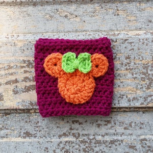 Puede incluir: Un posavasos cuadrado de ganchillo con un diseño de Mickey Mouse de color naranja brillante y verde. El posavasos está hecho con un hilo de color morado oscuro.