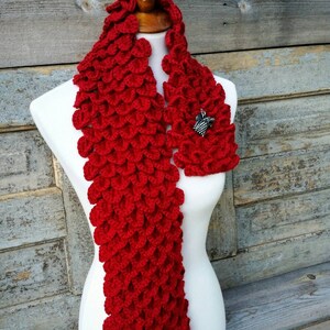 May include: Red crocheted scarf with a dragon scale pattern. The scarf is displayed on a white mannequin. A silver dragon pin is attached to the scarf.