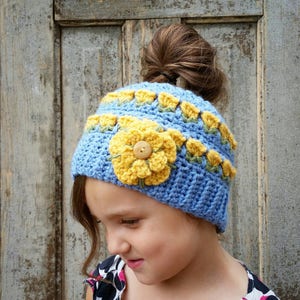 May include: A blue crocheted hat with yellow flower accents and a button in the center. The hat is designed to be worn with a ponytail.