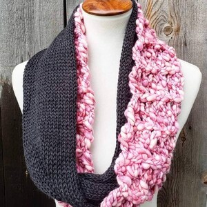 May include: A gray and pink knitted infinity scarf. The scarf is made with two different types of yarn, one is a smooth knit and the other is a chunky knit.