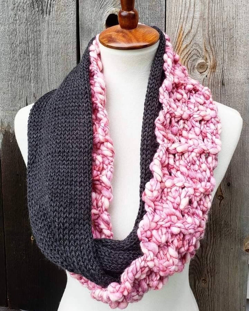 May include: A gray and pink knitted infinity scarf. The scarf is made with two different types of yarn, one is a smooth knit and the other is a chunky knit.
