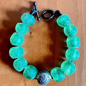 May include: A green glass bead bracelet with a silver clasp and a silver bead with a floral design.