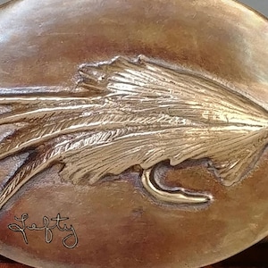 May include: A bronze belt buckle with a detailed fly design. The buckle is oval shaped and has a signature "Lefty" inscribed on the bottom.