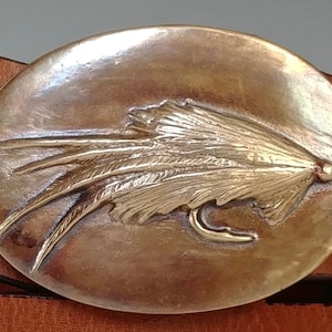 May include: A brass belt buckle with a detailed design of a fly fishing lure. The buckle is oval shaped and has a shiny finish.