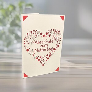 May include: A cream-colored Mother's Day card with a heart-shaped cutout design. The heart is filled with white flowers and red hearts, with the German text "Alles Gute zum Muttertag!" The card has red corner accents.