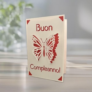 May include: A folded greeting card with a red and white butterfly design. The card has the Italian words "Buon Compleanno!" printed on the front. Red corner accents add a decorative touch. The card is on a light-colored surface.