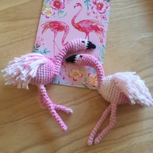 May include: Two pink crocheted flamingo toys with white feathers. The flamingo on the left has its head bent back and the flamingo on the right has its head bent forward.