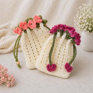 Puede incluir: Dos bolsas de crochet color crema con cordón y detalles florales. Las bolsas presentan un delicado tejido abierto y están adornadas con flores de ganchillo rosas y verdes alrededor de la parte superior. Los cordones verdes con cuentas de madera completan el diseño. Una bolsa tiene flores rosas, la otra fucsia.