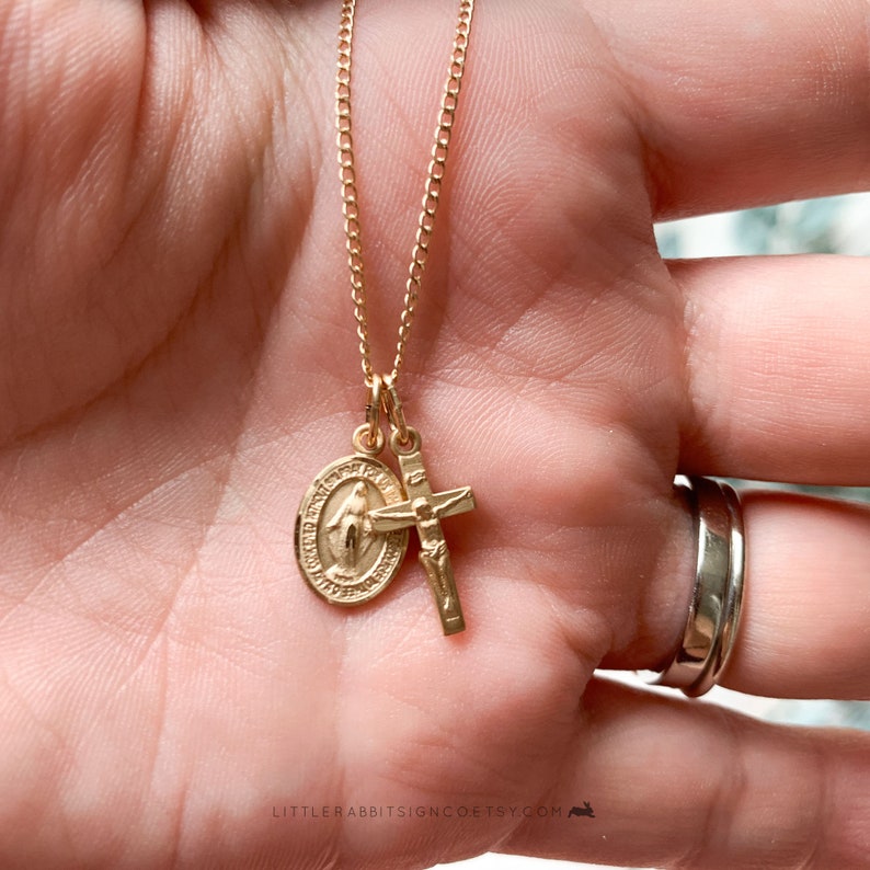 May include: Gold chain necklace with two charms: a round, detailed medal with a religious figure and a simple cross.