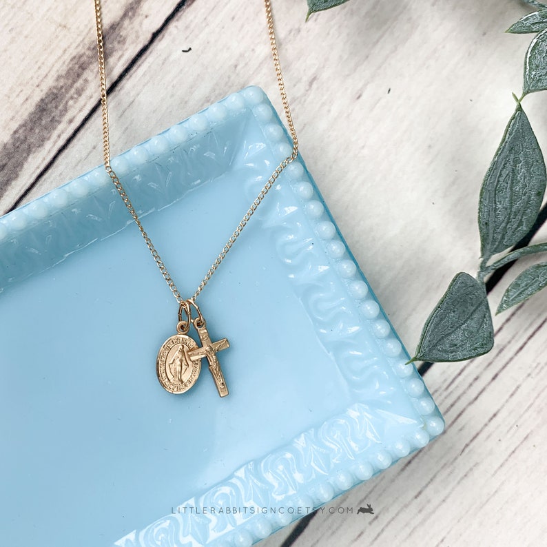May include: A gold chain necklace with a small gold cross and a gold medal charm featuring the Virgin Mary. The necklace is displayed on a light blue ceramic dish.