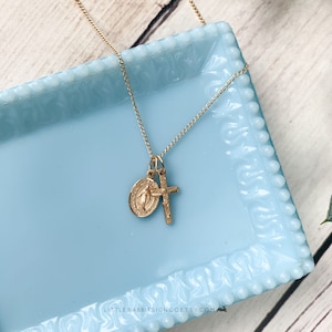 May include: A gold chain necklace with a small gold cross and a round gold pendant featuring a religious image. The necklace is displayed on a light blue tray.