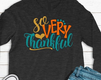May include: Dark gray long-sleeve shirt with the text "So Very Thankful" in orange, teal, and gold. The shirt is paired with blue jeans and white Converse sneakers, creating a casual, everyday look. The shirt is made of a soft material.